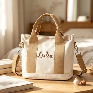 May include: A cream-colored canvas tote bag featuring two tan fabric handles and a matching detachable shoulder strap. The front of the bag displays the name Lidia embroidered in a brown script font. The bag has a structured rectangular base.