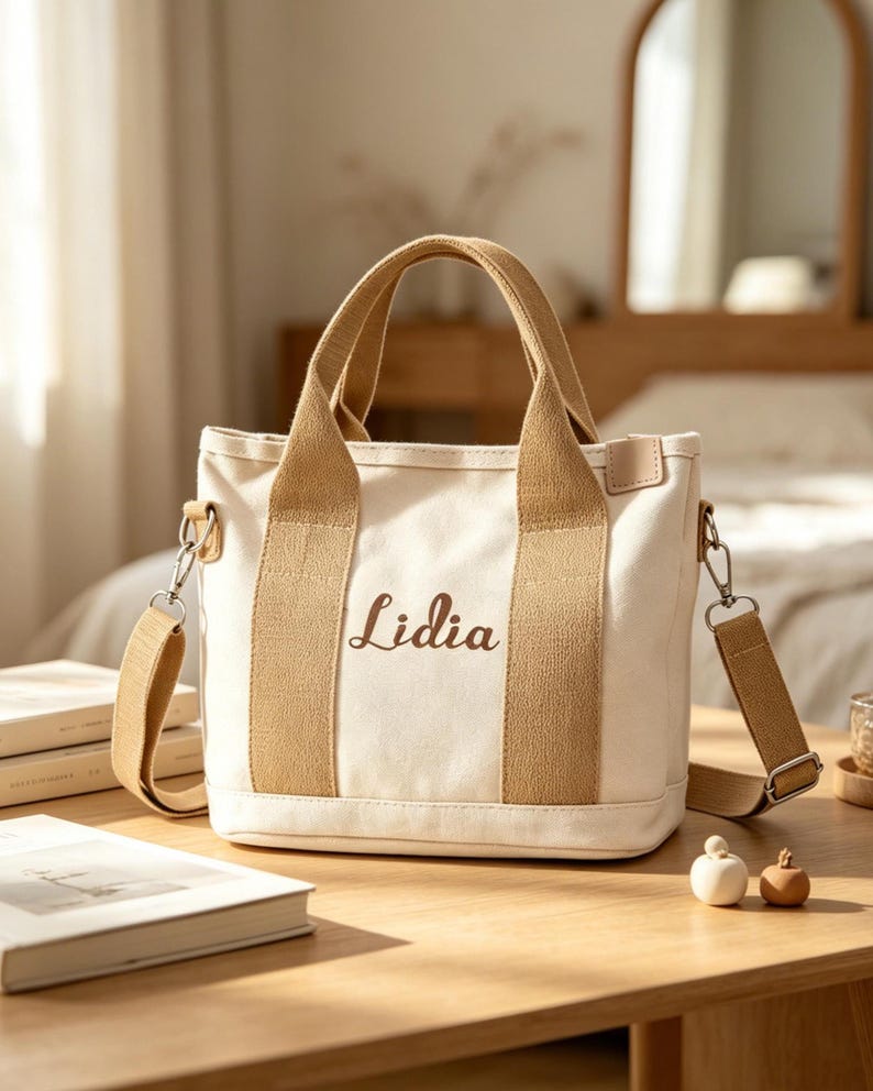 May include: A cream-colored canvas tote bag featuring two tan fabric handles and a matching detachable shoulder strap. The front of the bag displays the name Lidia embroidered in a brown script font. The bag has a structured rectangular base.