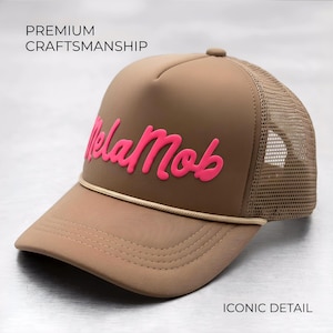 Puede incluir: Gorra de camionero color canela con parte trasera de malla y visera curva. La gorra presenta el texto rosa "Mela Mob" en la parte delantera. También son visibles las palabras "Premium Craftsmanship" e "Iconic Detail".