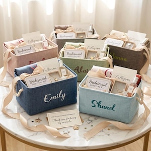 Op de afbeelding: Verschillende cadeau-manden in diverse kleuren, met touwhandvatten, gevuld met diverse items. De manden zijn voorzien van namen zoals "Emily" en "Shanel". Elke mand bevat cadeaus en kaarten. Op een tafel geplaatst.