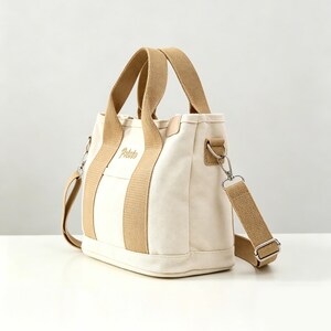 May include: A cream-colored canvas tote bag featuring tan webbing handles and a matching detachable shoulder strap. The front of the bag displays the word Potato embroidered in a tan script font. It includes silver-toned metal hardware.