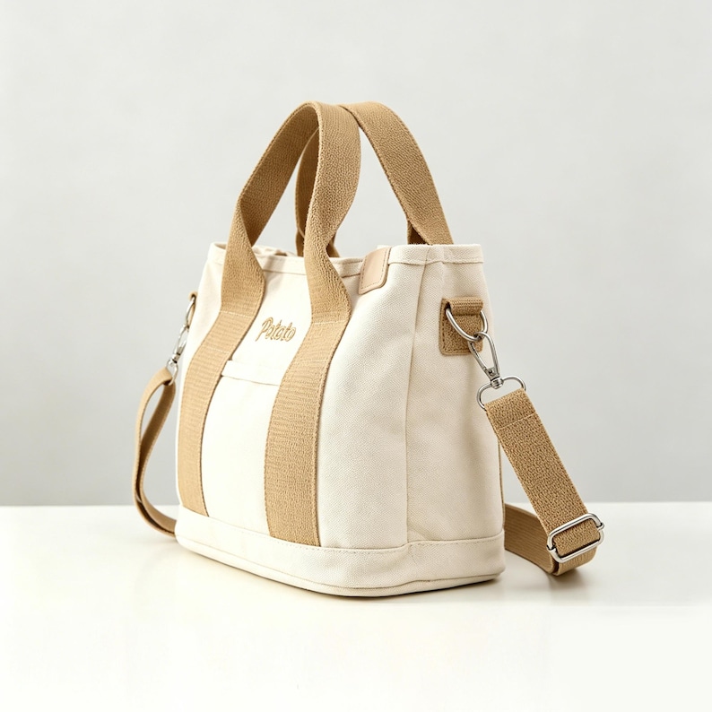 May include: A cream-colored canvas tote bag featuring tan webbing handles and a matching detachable shoulder strap. The front of the bag displays the word Potato embroidered in a tan script font. It includes silver-toned metal hardware.