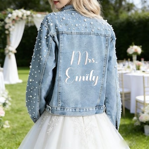 Op de afbeelding: Lichtblauwe spijkerjas versierd met imitatieparels. De achterkant van de jas toont de tekst "Mrs. Emily" in een sierlijk handschrift. De jas maakt deel uit van een bruidsensemble, ideaal voor een bruiloft.