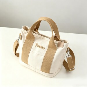 May include: A cream-colored canvas tote bag featuring tan fabric handles and a matching detachable shoulder strap. The front of the bag displays the word Potato embroidered in tan thread. It includes silver-toned metal hardware and clips.