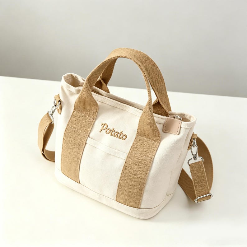 May include: A cream-colored canvas tote bag featuring tan fabric handles and a matching detachable shoulder strap. The front of the bag displays the word Potato embroidered in tan thread. It includes silver-toned metal hardware and clips.