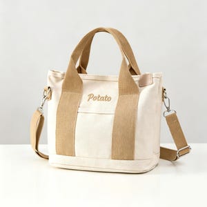 May include: A cream-colored canvas tote bag featuring tan fabric handles and a matching detachable shoulder strap. The word Potato is embroidered in brown script on the front center, positioned above a small horizontal pocket with silver hardware.