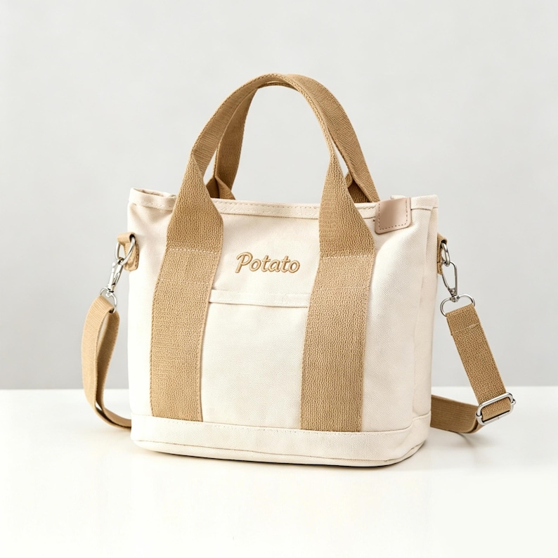 May include: A cream-colored canvas tote bag featuring tan fabric handles and a matching detachable shoulder strap. The word Potato is embroidered in brown script on the front center, positioned above a small horizontal pocket with silver hardware.