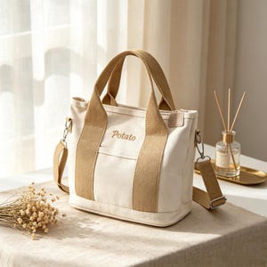 May include: A cream-colored canvas tote bag featuring tan fabric handles and a matching detachable shoulder strap. The word Potato is embroidered in brown thread on the front center. The bag includes a small exterior pocket and silver hardware.