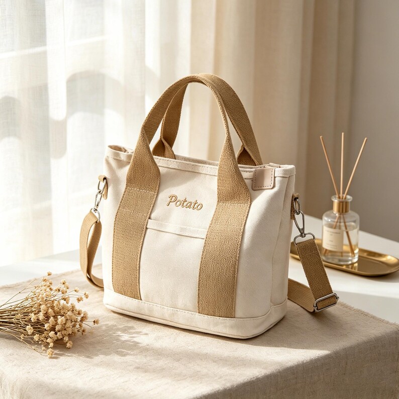 May include: A cream-colored canvas tote bag featuring tan fabric handles and a matching detachable shoulder strap. The word Potato is embroidered in brown thread on the front center. The bag includes a small exterior pocket and silver hardware.
