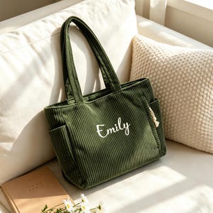 Op de afbeelding: Olijfgroene corduroy tote bag met twee zijzakken en stevige handvatten. De naam "Emily" is in witte letters op de voorkant geborduurd. De tas ligt op een witte bank met een beige kussen.