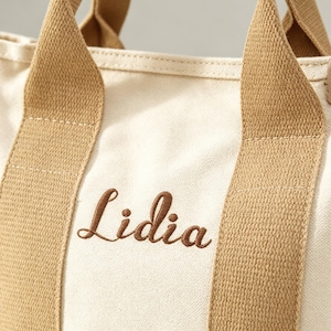 May include: A cream-colored canvas tote bag featuring two sturdy tan fabric handles and the name Lidia embroidered in brown script lettering on the front. The bag is constructed from a woven textile material with visible stitching along the straps.