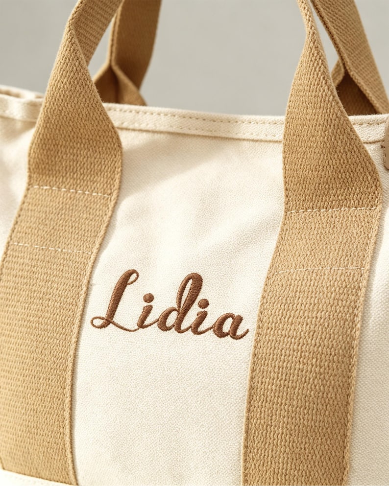 May include: A cream-colored canvas tote bag featuring two sturdy tan fabric handles and the name Lidia embroidered in brown script lettering on the front. The bag is constructed from a woven textile material with visible stitching along the straps.