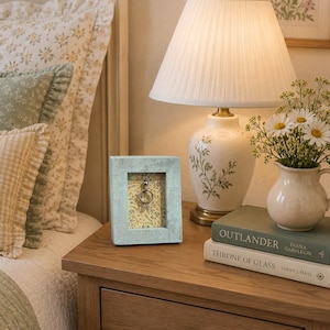May include: A light blue rectangular wooden frame with a distressed finish, containing a small silver ring suspended against a cream-colored background featuring a delicate floral pattern of small leaves and flowers in shades of green and yellow.