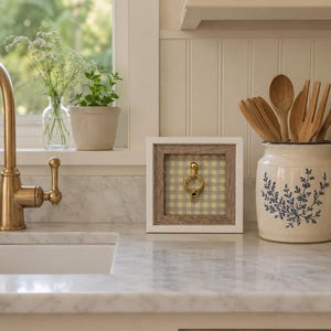May include: A square shadow box frame with a light wood inner border and a white outer frame. The background inside the frame features a yellow and white gingham pattern. A gold-toned metal ring knocker is mounted in the center of the frame.