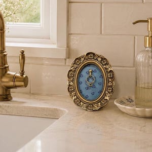 May include: An oval-shaped, antique-style brass ring holder frame featuring an ornate, embossed floral border. The center displays a light blue background with a small, repeating dark blue flower pattern and a central metal hook holding a ring.