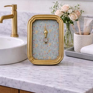 May include: A gold-colored framed key holder with a floral patterned background. The key holder is on a marble countertop in a bathroom setting. A gold faucet and a vase of flowers are also visible.