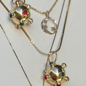 Pode incluir: Dois berloques esféricos dourados com pequenas pedras incrustadas em vermelho e verde alternados, suspensos em correntes finas de ouro ao lado de um pingente circular plano de ouro que retrata uma lua crescente e uma pequena estrela.