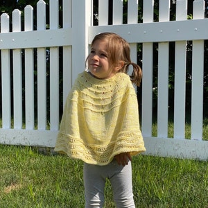 May include: A light yellow knitted poncho with a decorative pattern. The poncho has a round neckline and a flared bottom edge. The child is wearing gray pants and is standing in front of a white picket fence.