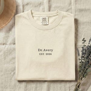 May include: Cream-colored t-shirt with the text "Dr. Avery EST. 2026" printed in black. The shirt is folded and lies on a neutral-toned surface. A straw hat and a bundle of dried flowers are also visible.