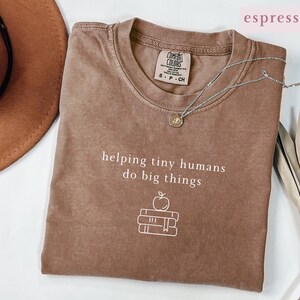 May include: An espresso-colored short-sleeve t-shirt featuring the white text 'helping tiny humans do big things' above a line-art graphic of an apple resting on a stack of three books. The shirt has a crew neckline and a Comfort Colors brand label.