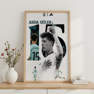 May include: A framed vertical poster featuring a collage of soccer player Arda Güler in a white jersey. The design includes the name ARDA GÜLER, the number 15, the Real Madrid crest, and the Adidas logo. The player is shown in various poses.