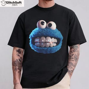 May include: Black t-shirt featuring a cartoon character with blue fur, large white eyes, and a mouth with diamond-studded teeth. The design is centered on the shirt. The GlitchSoft Store logo is in the top left corner.