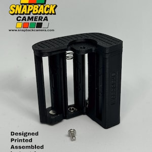 May include: A black 3D-printed battery holder for a Snapback Camera with visible metal springs and contacts. The unit features a textured top surface and includes a small silver metal screw. Text reads: SNAPBACK CAMERA, www.snapbackcamera.com, Designed Printed Assembled in Austria.