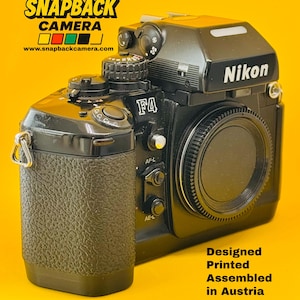 May include: A black Nikon F4 film camera with a textured grip, a lens cap, and various control dials. The text SNAPBACK CAMERA and www.snapbackcamera.com appear at the top, while Designed Printed Assembled in Austria is printed at the bottom.