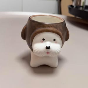 Pode incluir: Um vaso de cerâmica em forma de cabeça de cão com cara branca, nariz preto e detalhes cor-de-rosa nas bochechas. O cão usa um capuz castanho texturizado que forma a borda do recipiente. O interior do vaso é de cor creme e oco.