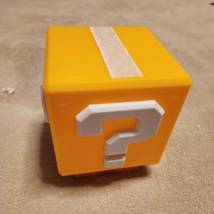 May include: A yellow cube featuring a white question mark, possibly a toy or prop. A white stripe runs across the top. The design is inspired by a popular video game.