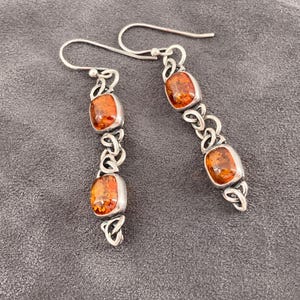 Könnte beinhalten: Ein Paar silberner Ohrhänger mit rechteckigen, bernsteinfarbenen Steinen. Die Ohrringe haben ein keltisches Knotendesign zwischen den Steinen und am unteren Ende. Die Ohrringe werden auf einer grauen Oberfläche präsentiert.