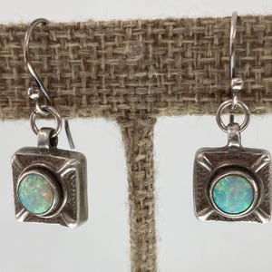 May include: A pair of silver-toned earrings with a square design. Each earring features a round, iridescent opal stone set in the center. The earrings are displayed on a neutral-colored surface.