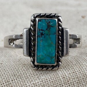 Puede incluir: Un anillo de plata con una piedra rectangular de turquesa. La piedra es de color turquesa vibrante con vetas oscuras. El anillo tiene un diseño de cuerda trenzada alrededor de la piedra y elementos decorativos en los laterales.