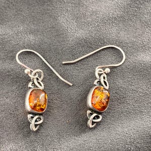 May include: Silver earrings featuring amber-coloured, rectangular stones. The earrings have a Celtic knot design and a hook. The amber stones display a warm, golden-orange hue. The earrings are a unique piece of jewellery.