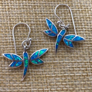 May include: A pair of silver dragonfly earrings with blue and green iridescent wings. The earrings have a hook closure and are displayed on a textured, tan surface. The dragonfly design is detailed and eye-catching.