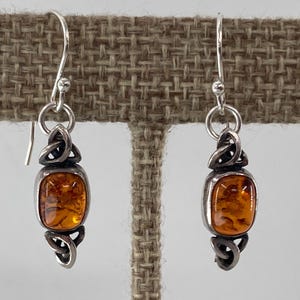 May include: Silver dangle earrings with amber-colored, square-shaped stones. The earrings feature a Celtic knot design above and below the amber stones. The hooks are silver.