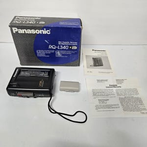 May include: A black Panasonic RQ-L340K mini cassette recorder with a black wrist strap, a gray battery cover, the original blue and white product box, an instruction manual, and a printed limited warranty document on a white surface.
