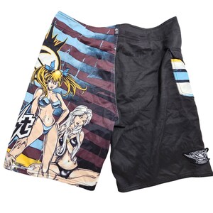 May include: Black and patterned board shorts. The left side features a graphic of two women in bikinis, with a sun and text. The right side is solid black with a pocket and a logo patch.