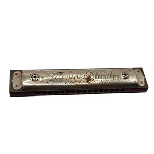 May include: A vintage harmonica with a dark brown body and a weathered silver-tone metal cover plate. The plate features the engraved text Zigeuner Musik and Made in Germany, with visible signs of oxidation and rust across the metallic surface.