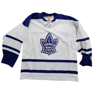 May include: White and blue Toronto Maple Leafs hockey jersey. The jersey features the team's logo on the front, with blue stripes on the sleeves and bottom. The collar and shoulders are blue.