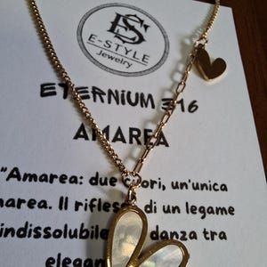 Può includere: Collana dorata con pendente a forma di cuore. Il pendente ha un intarsio in madreperla ed è sospeso a una catena sottile. Un piccolo ciondolo a forma di cuore è presente. La collana è esposta su una carta con testo.