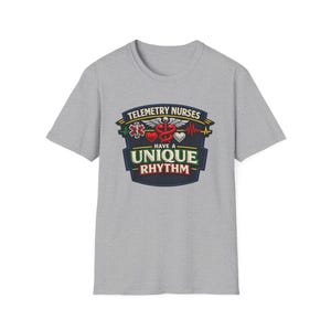 Könnte beinhalten: Ein graues, kurzärmeliges T-Shirt mit einem grafischen Aufdruck, der den Text TELEMETRY NURSES HAVE A UNIQUE RHYTHM enthält. Das Design zeigt einen medizinischen Caduceus, ein rotes Herz, ein weißes Herz und eine EKG-Linie auf einem dunkelblauen Schild.