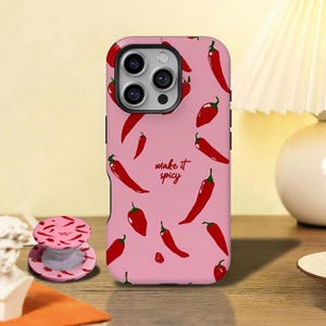 Puede incluir: Funda de teléfono rosa y agarre a juego con pimientos rojos. La funda del teléfono tiene la frase "make it spicy" en fuente manuscrita. El agarre del teléfono es redondo y tiene un diseño similar de pimientos.