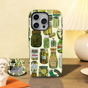 Peut inclure: Coque de téléphone avec un motif répété de bocaux de cornichons. Les bocaux, de formes et tailles variées, sont remplis de cornichons et étiquetés. La coque est vert clair, avec un pop socket assorti.