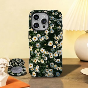May include: A smartphone case and a matching circular phone grip featuring a dense, repeating pattern of white daisy flowers with yellow centers set against a dark green, leafy background. The case has a cutout for a triple-lens camera system.