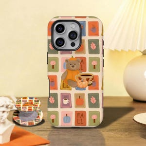 May include: A phone case and matching circular phone grip featuring a grid pattern of autumn-themed icons including a teddy bear wearing a pumpkin costume, pumpkins, coffee mugs, mushrooms, and leaves, with the text FALL in orange blocks.