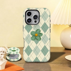 Könnte beinhalten: Eine Handyhülle mit grünem Rautenmuster und einer 3D-Blume in Grün. Ein passender Handyhalter ist ebenfalls zu sehen. Die Hülle liegt auf einer Holzoberfläche. Die Handyhülle schützt das Telefon vor Kratzern und Beschädigungen.