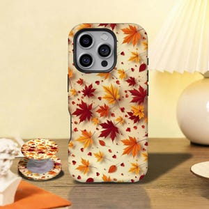 May include: A phone case and matching phone grip featuring a fall leaves design in shades of red, orange, and yellow. The case has a cream-colored background with scattered leaves and small hearts. The phone grip is round and has a similar design.