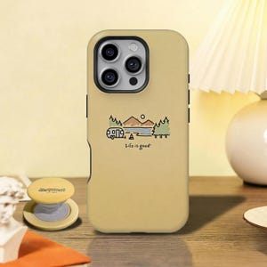 Op de afbeelding: Mosterdkleurige telefoonhoes met een kampeerthema en de tekst "Life is good!". De hoes heeft een ontwerp van een camper, bergen, bomen en een meer. Een bijpassende telefoonhouder is ook zichtbaar.