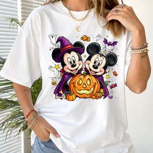 May include: A white t-shirt featuring a graphic of two cartoon mice dressed in purple witch and vampire costumes, standing behind a large orange jack-o'-lantern. The design includes small ghosts, spiderwebs, stars, bones, and colorful candies.